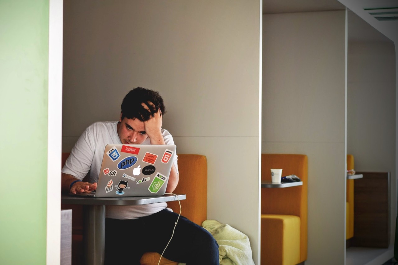 A student in a white shirt sits in front of a laptop with his hand on his head.