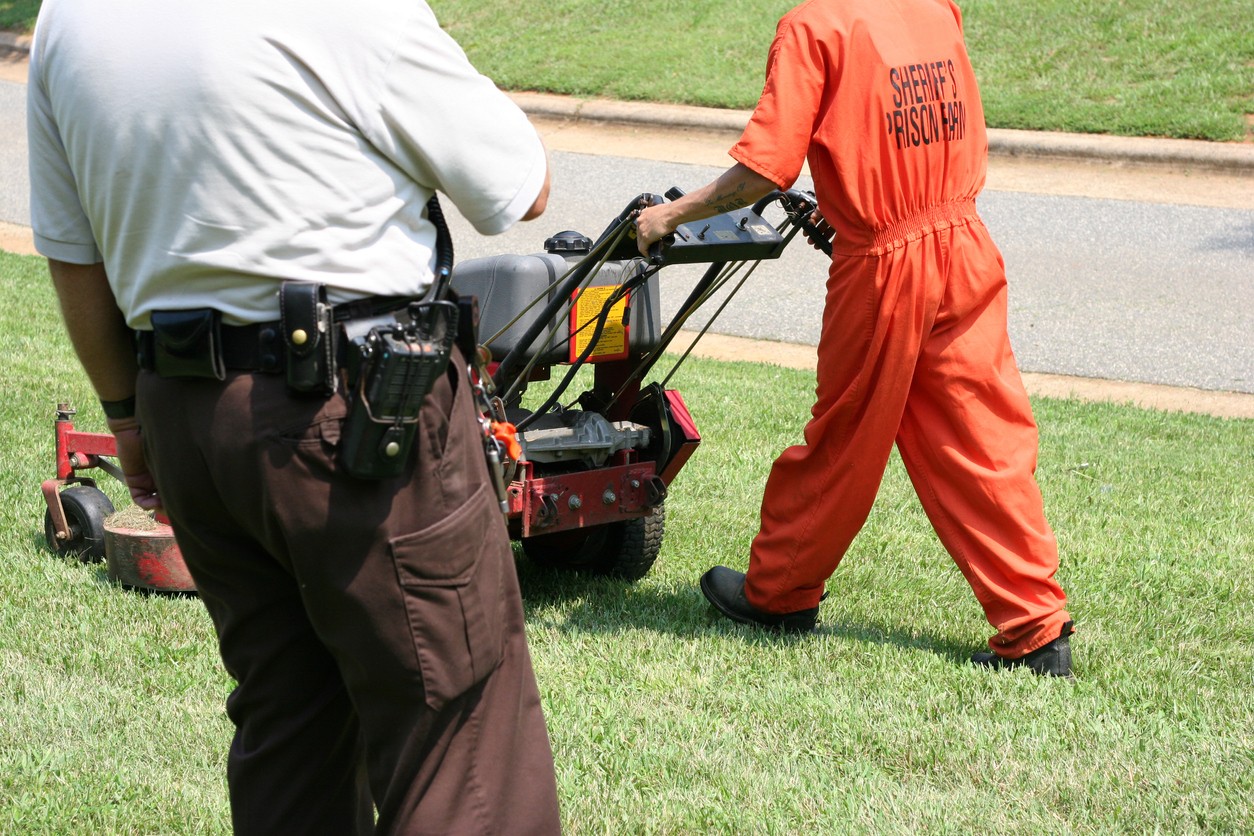 Prison worker cutting grass while guard watches
