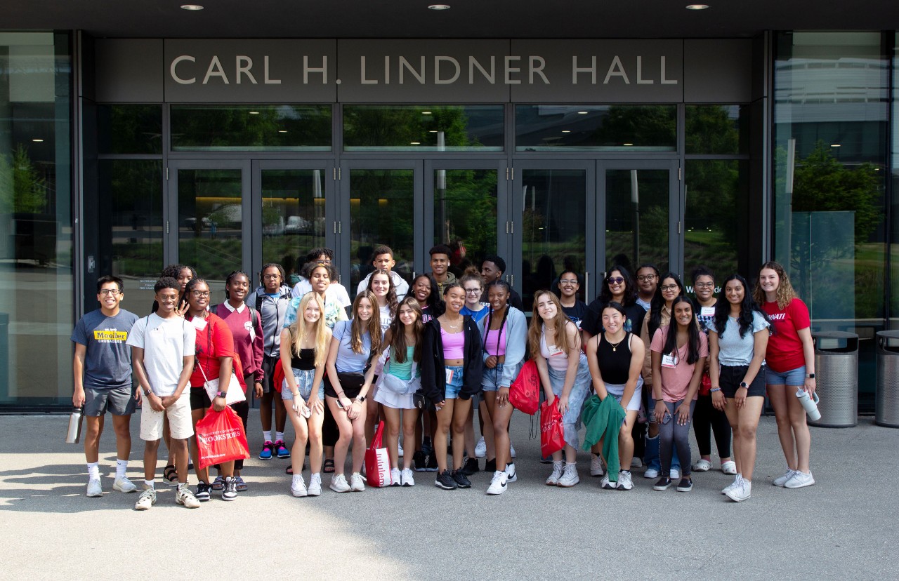 A group of 30 high school students stand in front of Lindner Hall in morning light wearing multi color shorts and T-shirts.