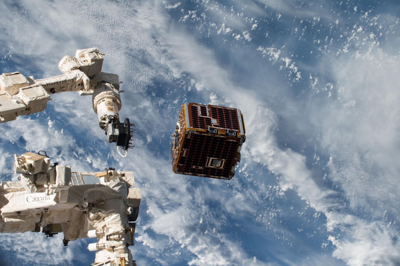 A robotic arm releases a satellite into orbit over the Earth. A vast ocean is visible through the clouds.