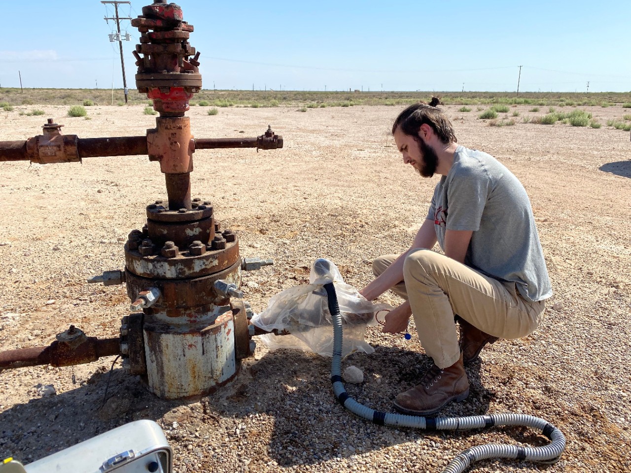 A UC student kneels in a desert next to rusty piping sticking out of the ground and holds electronic equipment.