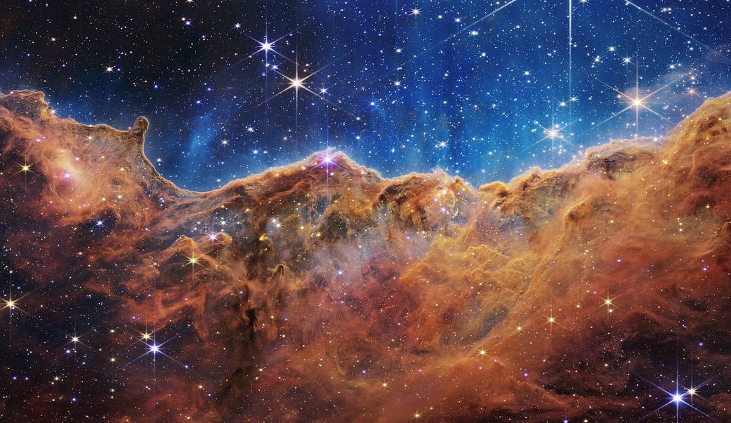 The James Webb Space Telescope captures an image of the Carina Nebula showing a red, pink and orange cloudlike formation in front of a field of stars and a blue background.