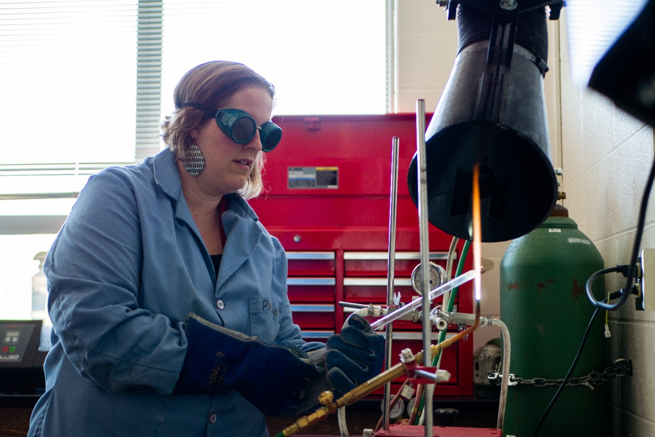 Woman in a blue lab coat, protective goggles and gloves holds metal over a flame in a lab