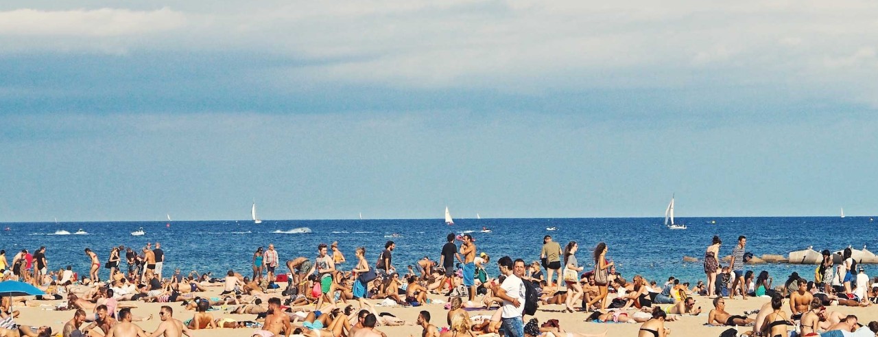 crowded beach with sun shining and sailboats in the background 