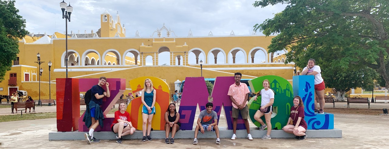 Young people pose, sitting and leaning, on colorful, free-standing letters that form a large sign reading “Izamal.” Behind them, a yellow and white wall with many archways separates the plaza from a monastary courtyard, with the bell towers of the yellow, Spanish-style church visible above it.