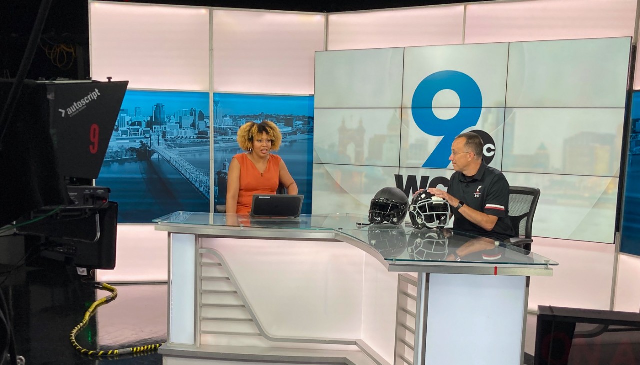 UC Professor Eric Nauman talks to WCPO reporter Kristen Swilley with two football helmets in front of him in the news studio.