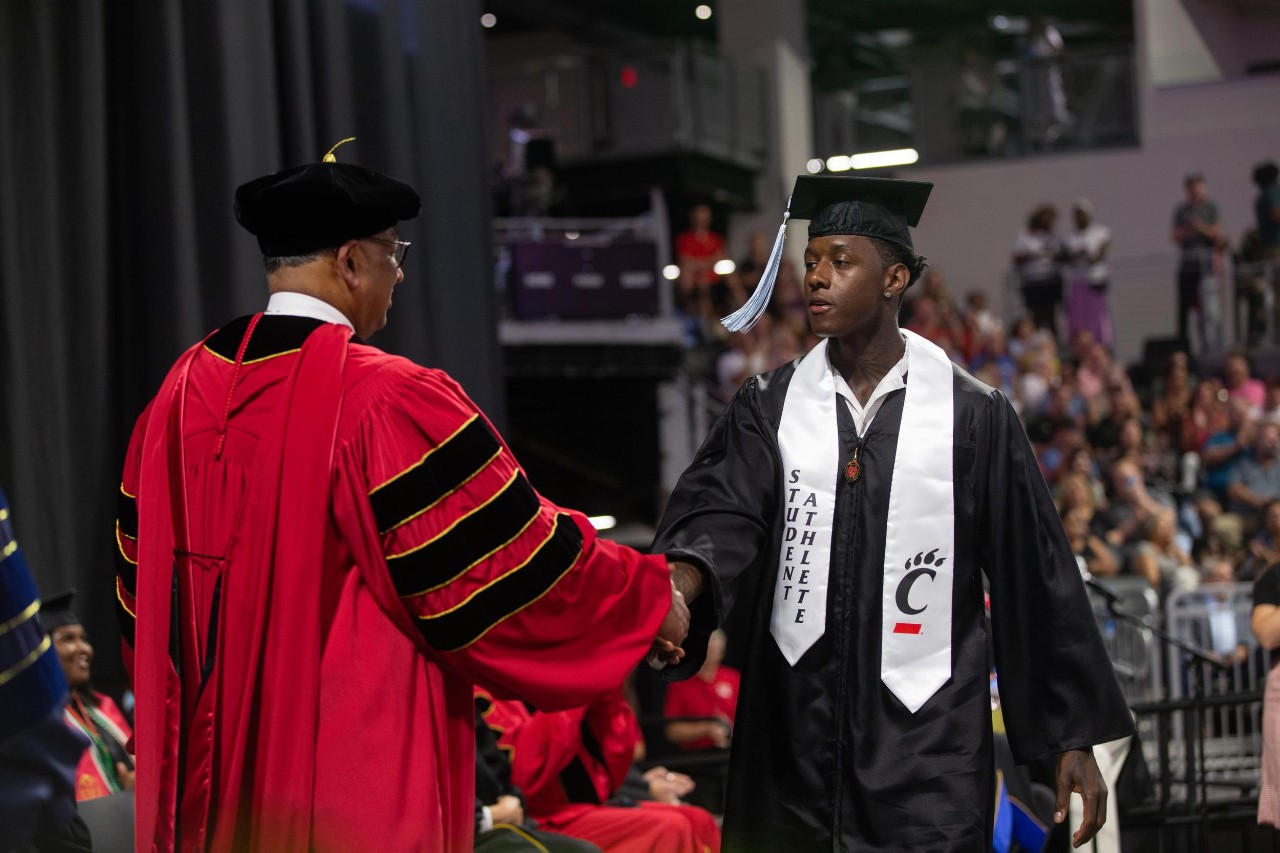 Ahmad "Sauce" Gardner in a cap and gown shakes hands with the UC president on stage.