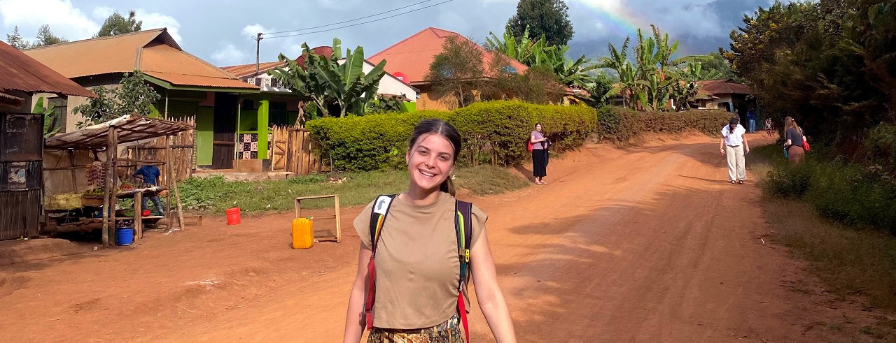 a young woman wearing a backpack stands smiling on a dirt road with small buildings and lush trees behind her; a rainbow is visible in the distance