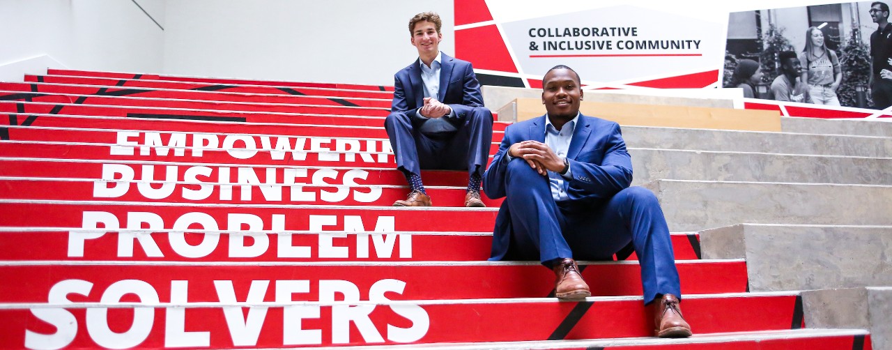 UC business students Patrick McNamara and Sharif Johnson sit on printed steps that state "Empowering Business Problem Solvers" inside the Lindner College of Business.