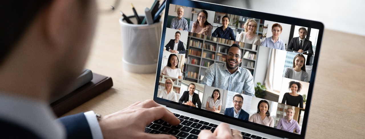 A businessman sits in front of a laptop on a virtual meeting with several boxes containing images of his colleagues