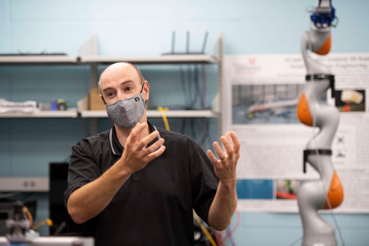 Andrew Barth wearing a face mask gestures in a lab surrounded by robotic arms.