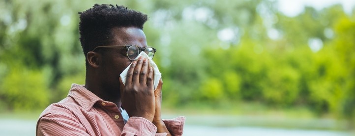 A man sneezes into a tissue in front of a lake and trees