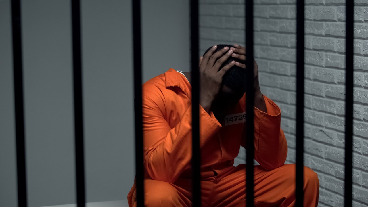 Prisoner sits in jail cell with his head in his hands. 