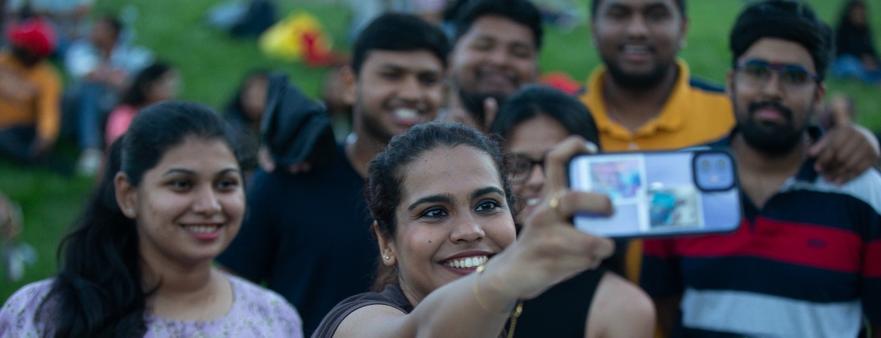 Seven international students from India posse for a selfie photo on the UC campus. Woman holds phone and everyone smiles.