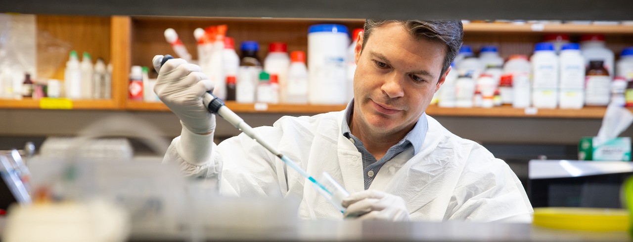 Tim Phoenix, wearing a white coat, uses a pipette to inject a sample into a test tube in his laboratory