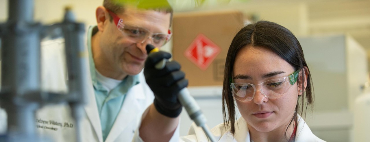 Research assistant Grace Goodhart sits at a bench and pipettes samples in a laboratory while Andrew Waters looks on. Both are wearing white laboratory coats.