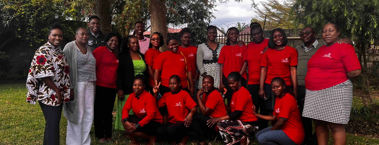 Leeya Pinder stands and smiles with men and women in front of a tree in Zambia. Some of the women are wearing red University of Cincinnati shirts.