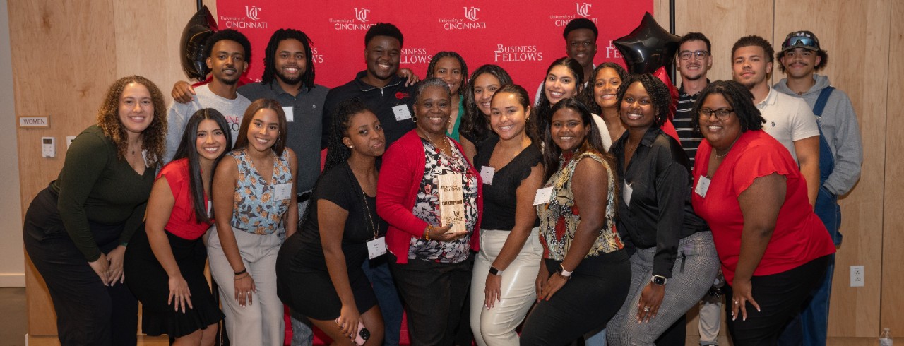 A group of 19 students pose with Kimberly McGinnis (center) in front of a red backdrop against a wall in the Lindner Hall atrium