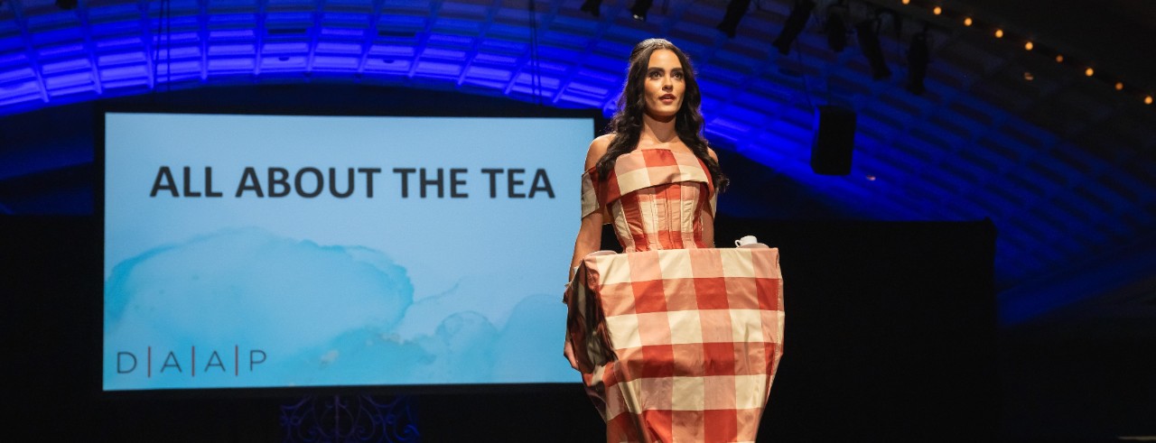 model on runway wearing a dress made out of a red and white tablecloth. The skirt of the dress is also a table with tea cups.