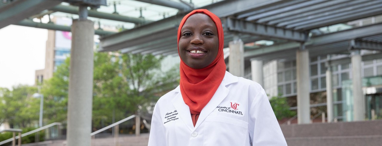 Graduate student Tasnim Olatoke is dressed in a white lab coat and standing on the steps of the UC College of Medicine.