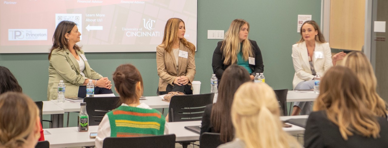 A panel of four women facing the camera at the front of a Lindner Hall classroom present to a classroom full of female business students