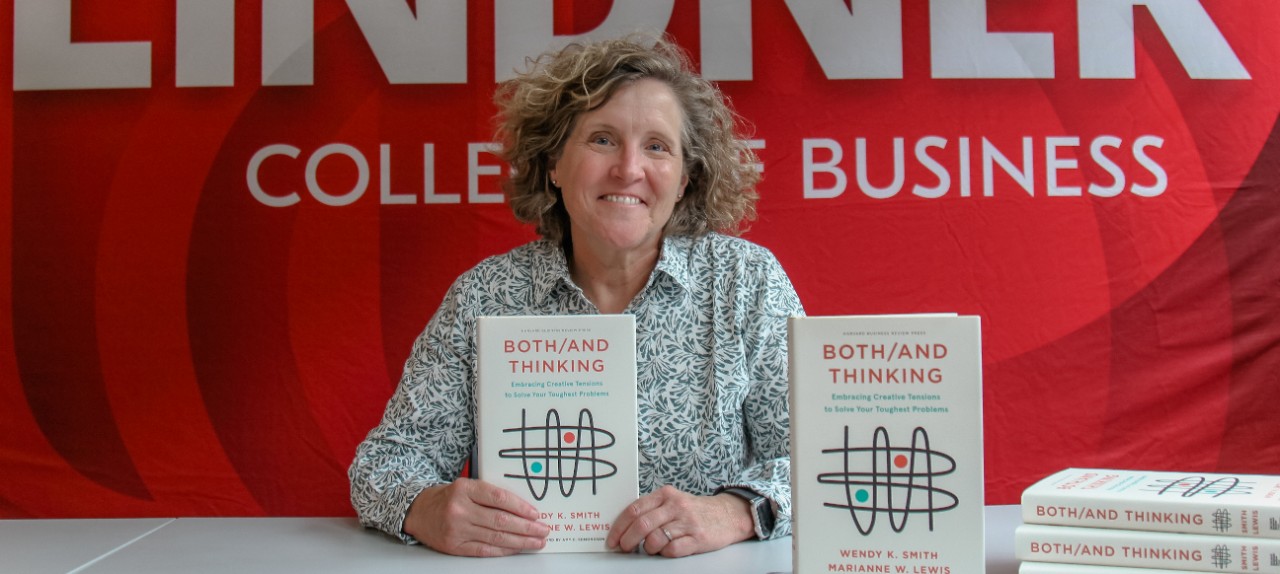 Dean Marianne Lewis holds up her book Both/And Thinking while sitting at a table in front of a Lindner College of Business banner.