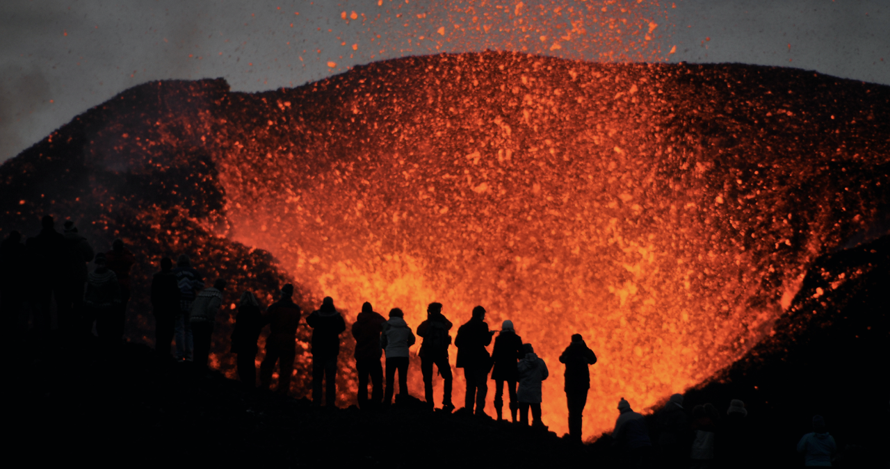 A row of people stand in the foreground as a volcano erupts in front of them at night.