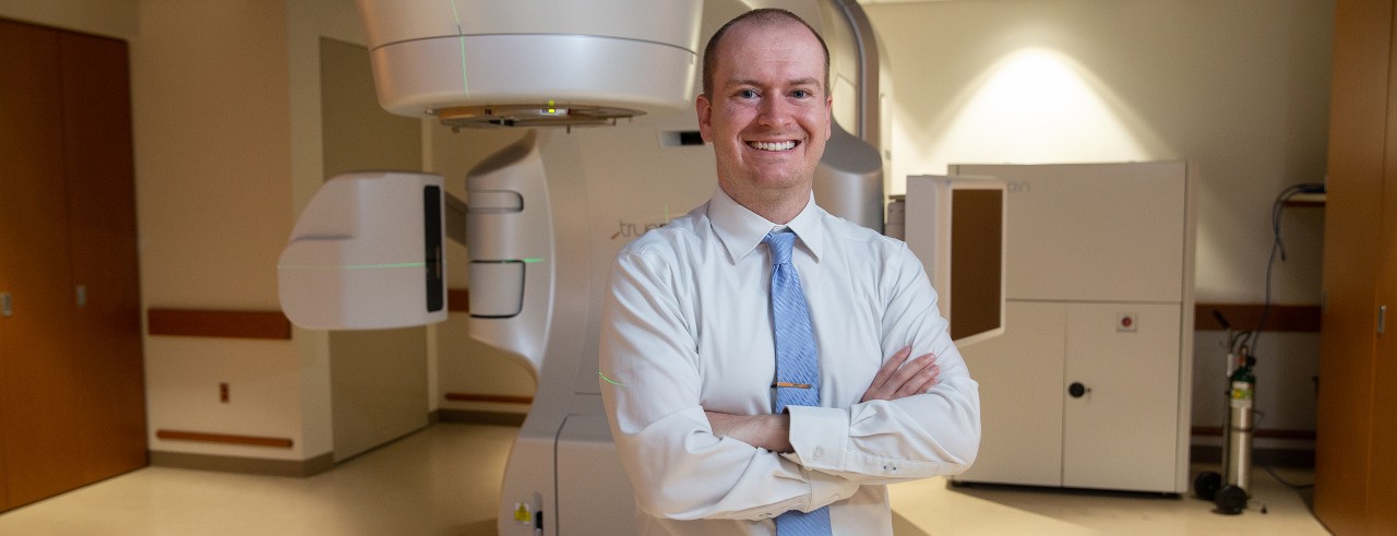 Andrew Frankart smiles standing in front of a radiation treatment machine
