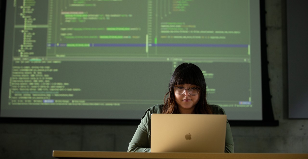 Female University of Cincinnati student works on laptop with a large screen of computer code in background.