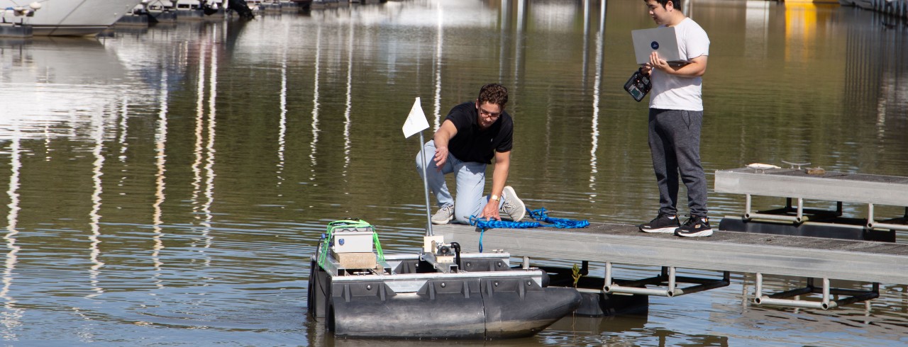 A man kneeling on a dock works on a rover in the water of a marina while another man with a laptop looks on.