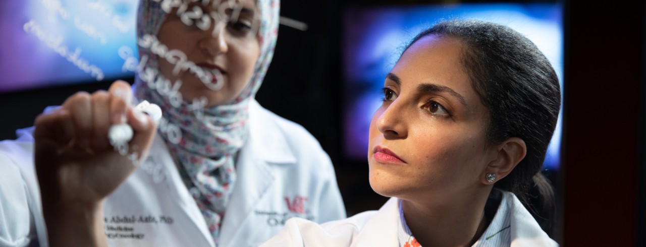 Ola Elgamal, wearing a white coat, writes an equation on a whiteboard while another researcher looks on