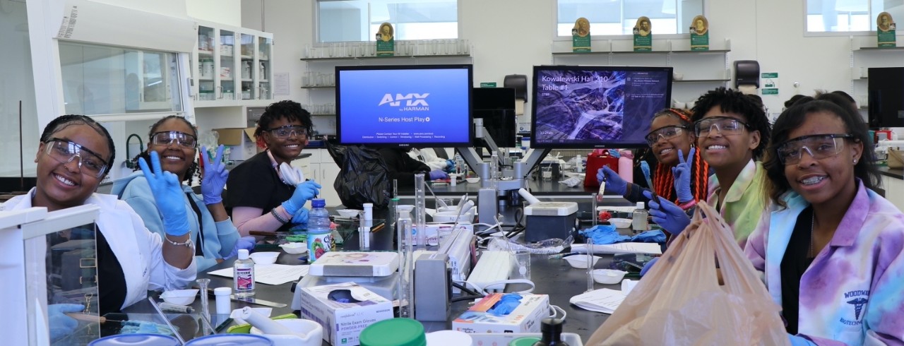 High school students wearing lab coats and safety glasses smile while working in a laboratory at the UC James L. Winkle College of Pharmacy