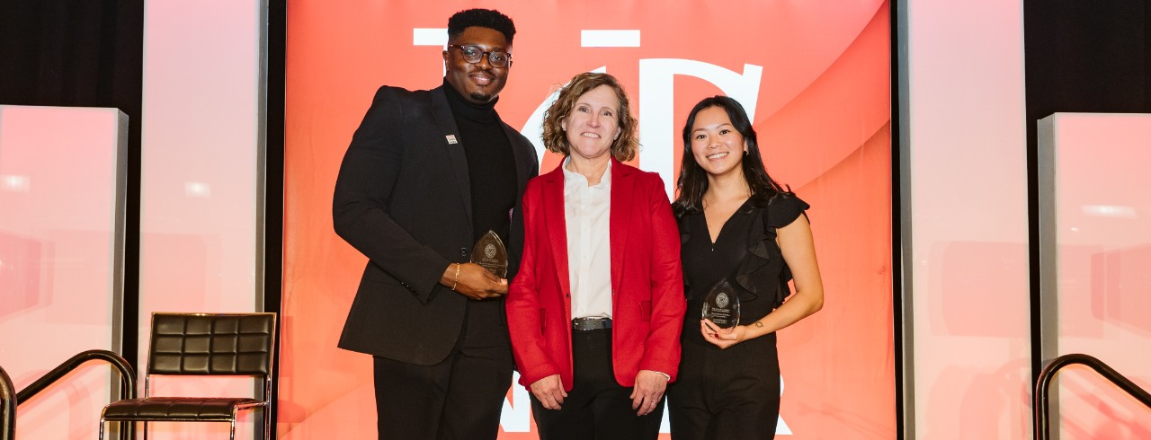 Three individuals stand in front of a red screen background. One man dressed in black holding a glass award on the left, one woman wearing red in the center and one woman wearing black holding a glass award on the right.