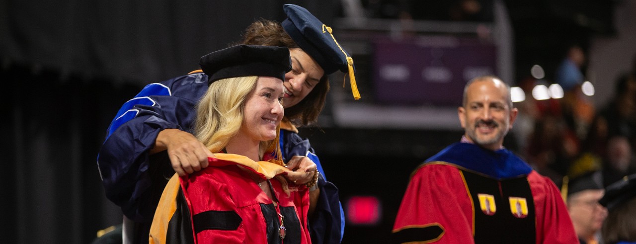 A doctoral student in a cap and gown is hooded on stage at Fifth Third Arena.