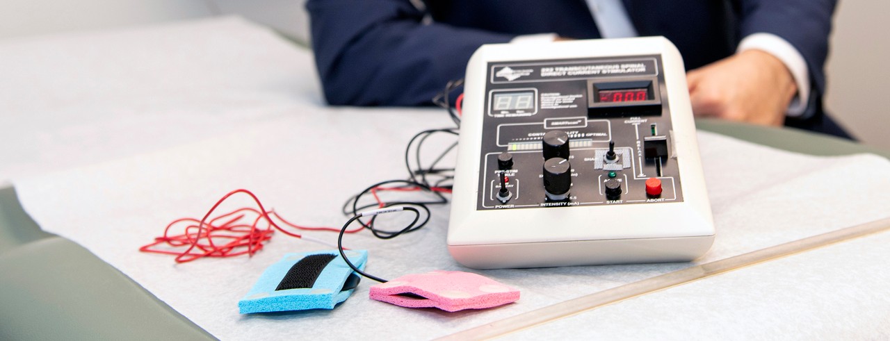 Blue and pink electrodes are connected by red and black wires to the spinal cord stimulation device, which has several knobs and switches