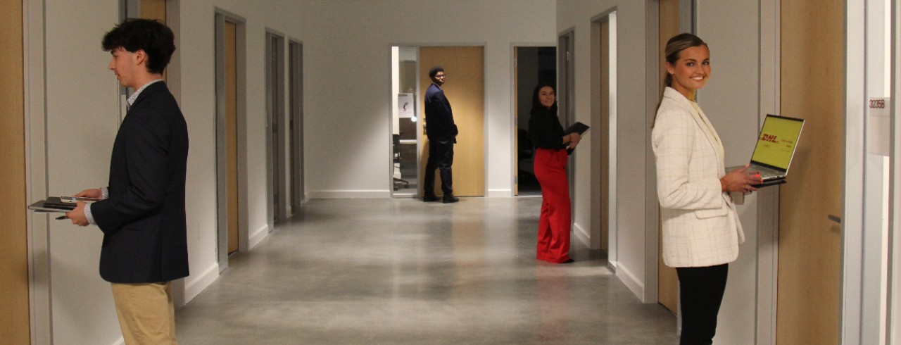 Four Lindner students stand in front of conference room doors, holding laptops