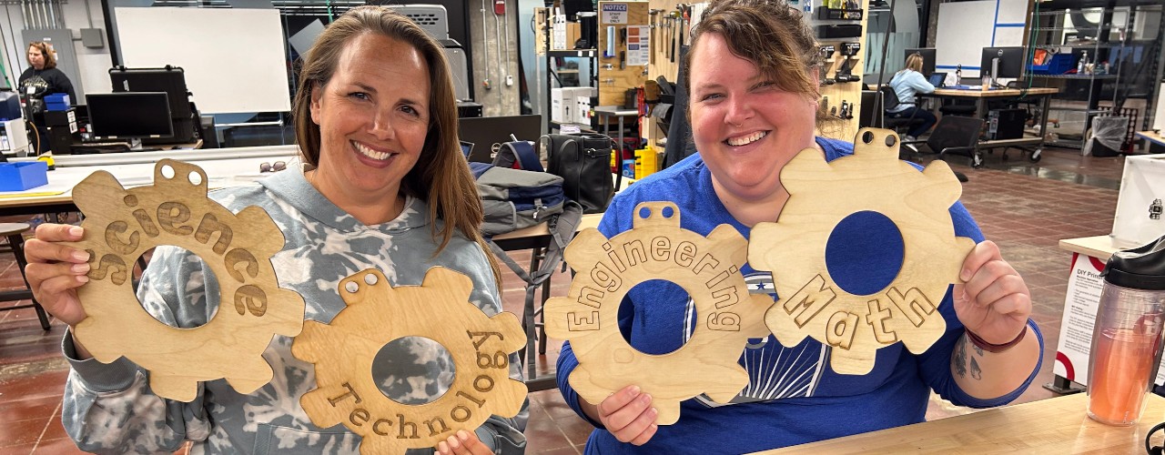 Two women hold stenciled gears that spell "science, technology, engineering, math."in UC's Makerspace