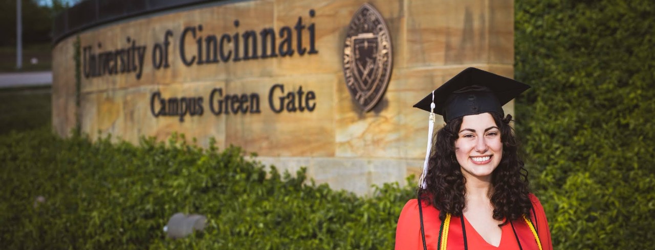 Trinity Shaya stands in her graduation cap and gown in front of a University of Cincinnati sign.