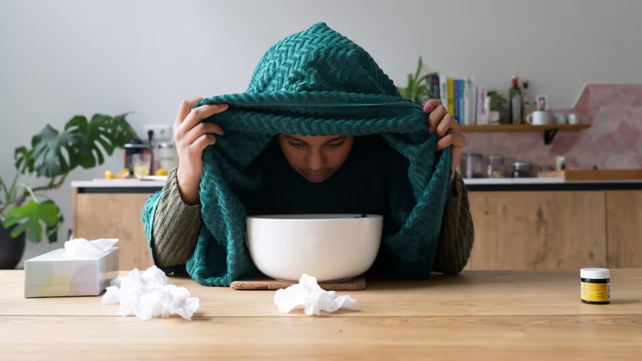 A person sitting at a table with their face over a bowl of steaming water holding a towel over their head