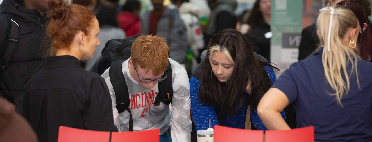 The backs of two housing agents are shown. Two students, a male and female, stand in between the agents and are shown signing sheets at the UC Off-Campus Housing Fair