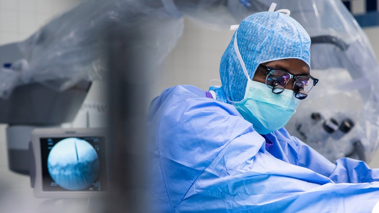 Dr. Adogwa wears blue scrubs, a scrub cab, mask and glasses while working in the operating room