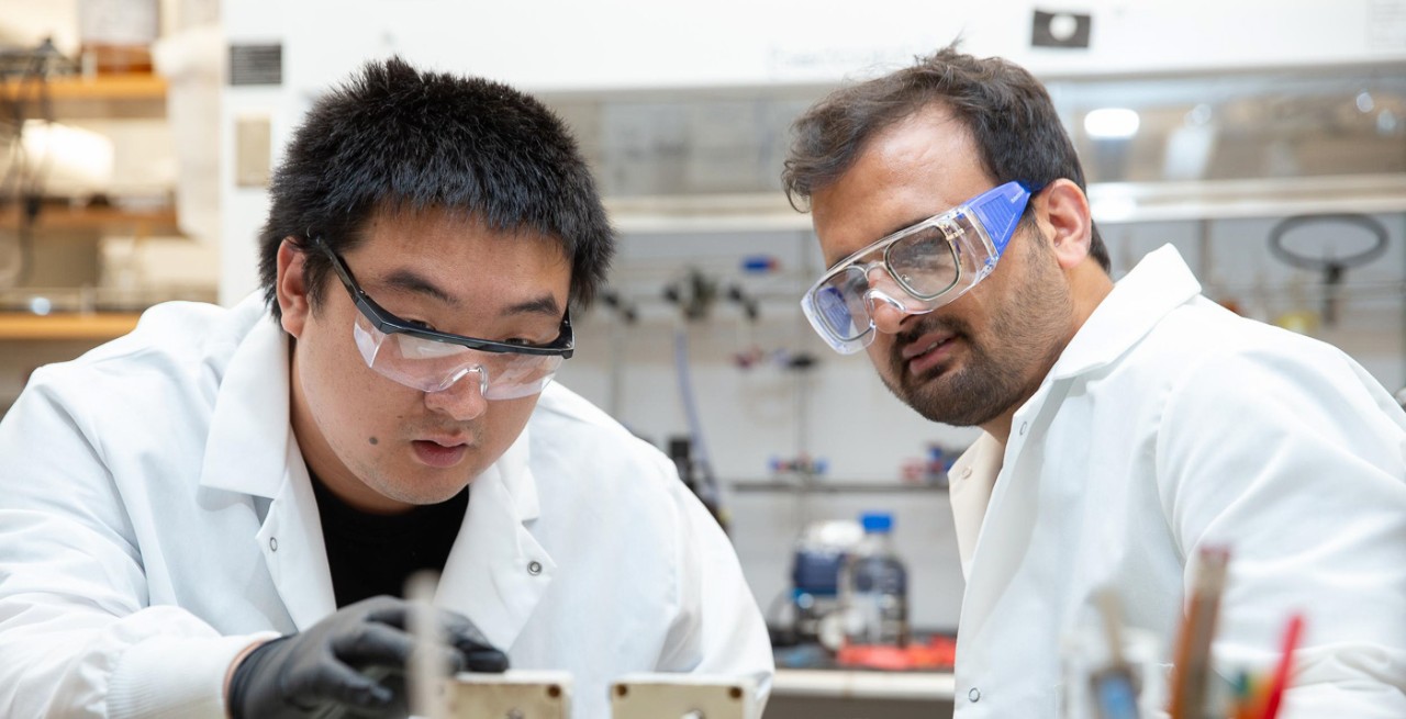 Doctoral students wearing labcoats and safety glasses work in a chemistry lab.