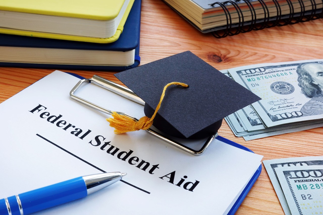 photo of a desk top showing a graduation hat on top of a pad with the word Federal Student Aid also two stacks of dollar bills nearby along with bookends shown