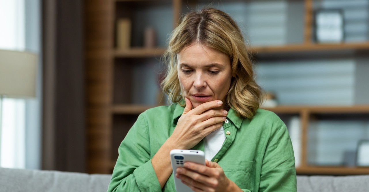 woman sitting on the couch wondering what number to call on her cell phone 
