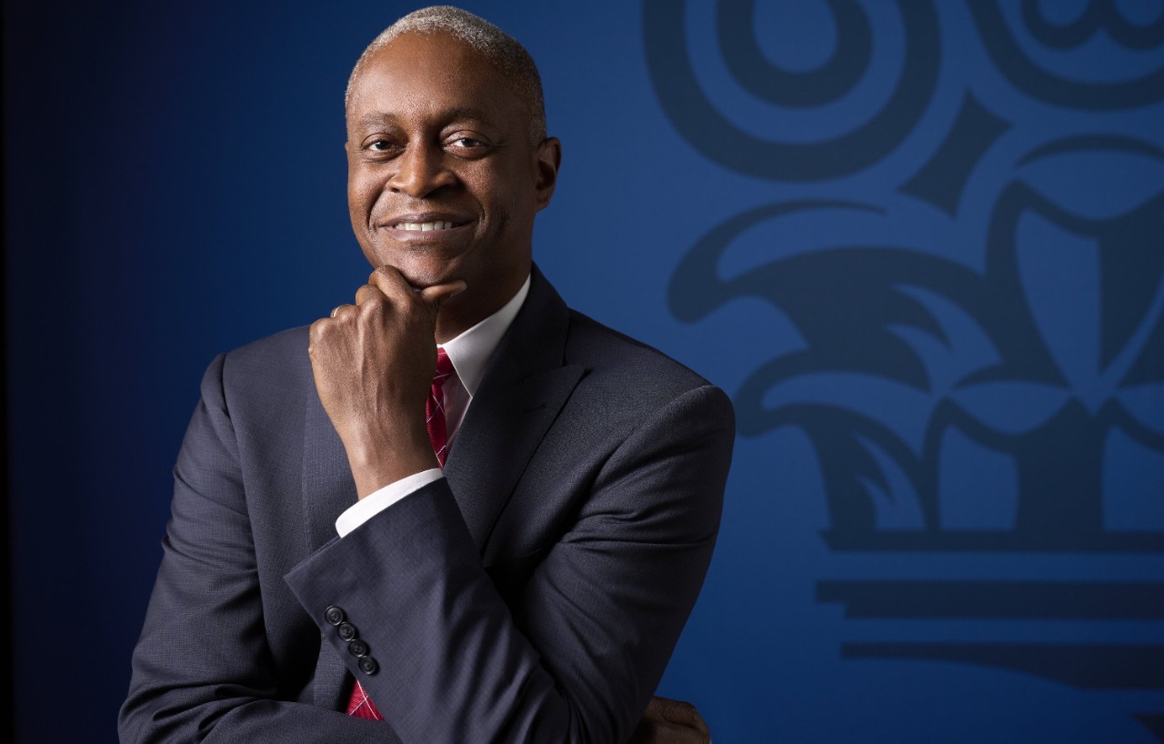 Raphael Bostic in a dark suit and red tie in front of a blue background.