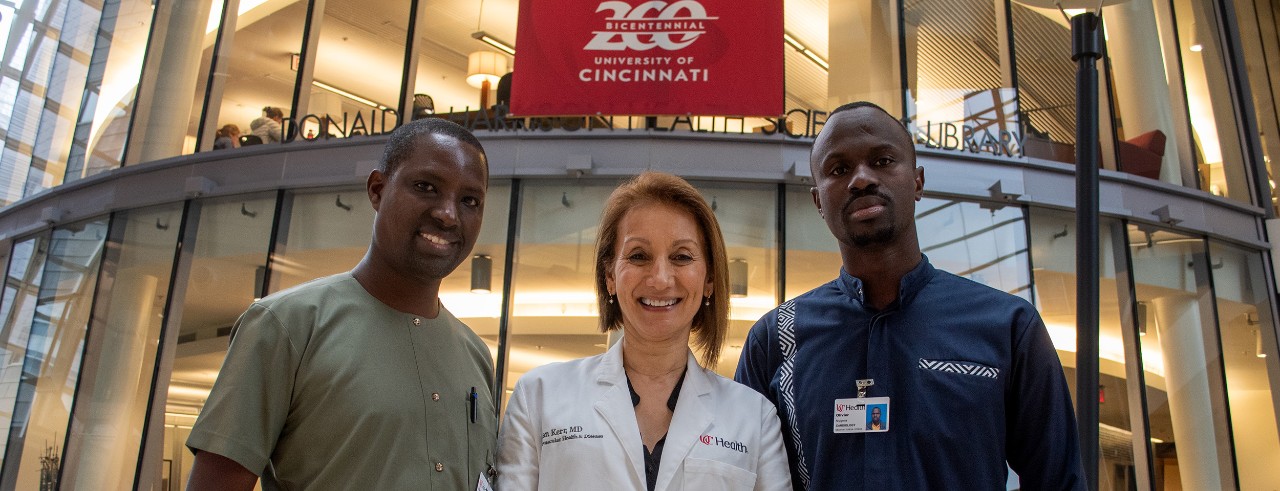 Bienvenu MUVUNYI, Hanan KERR and Olivier NIYIGENA stand, wearing UC Health scrubs or lab coats, stand in front of the Donald C. Harrison Health Sciences library in the University of Cincinnati College of Medicine before a red banner that reads," Bicentennial 200, University of Cincinnati."