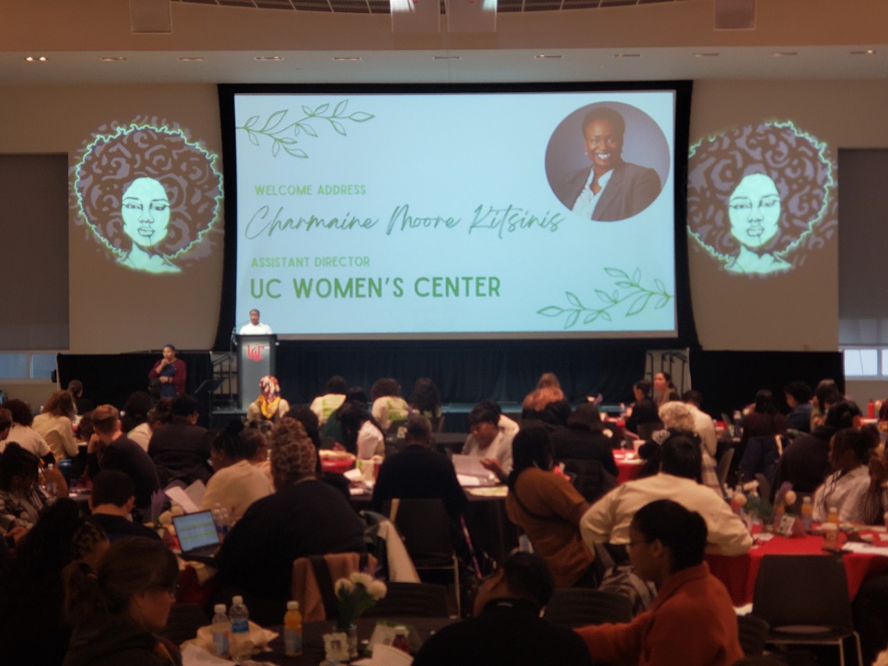 Image of Great Hall with hundreds of participants sitting at tables and listening to a woman speak at the Black Feminist Symposium. 