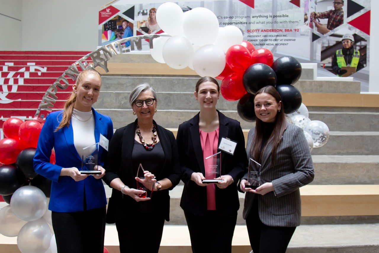Four women professional dressed stand in front of balloon backdrop in Lindner atrium holding their awards and smiling