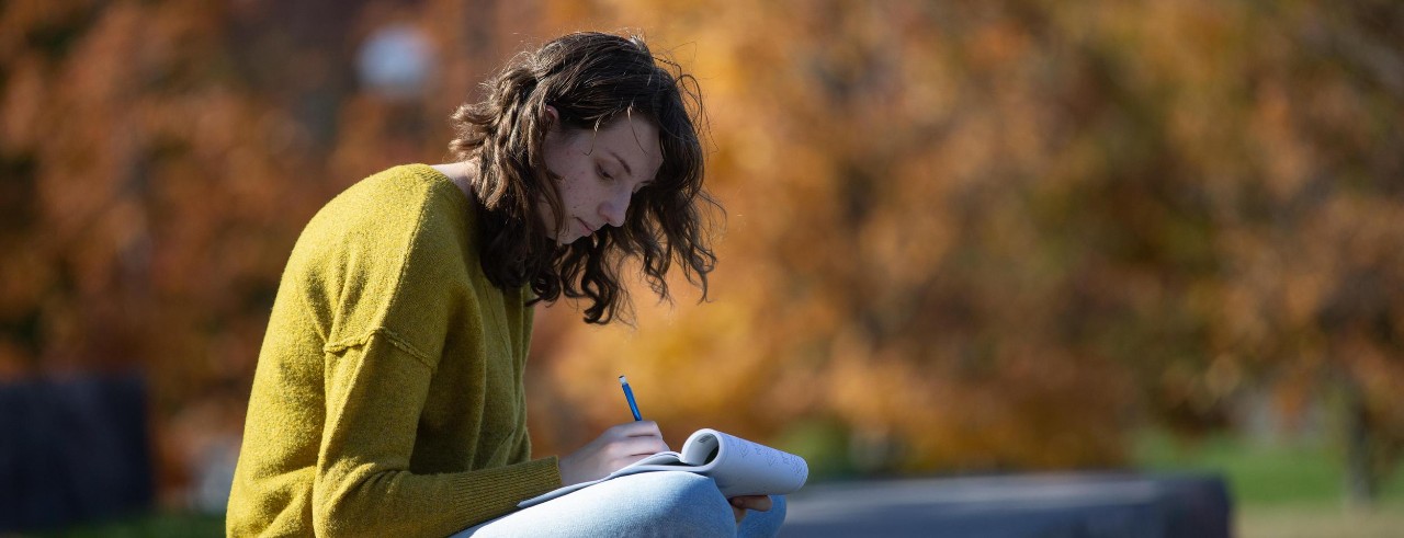 Female student sits on campus with a notepad writing. Trees in the background.