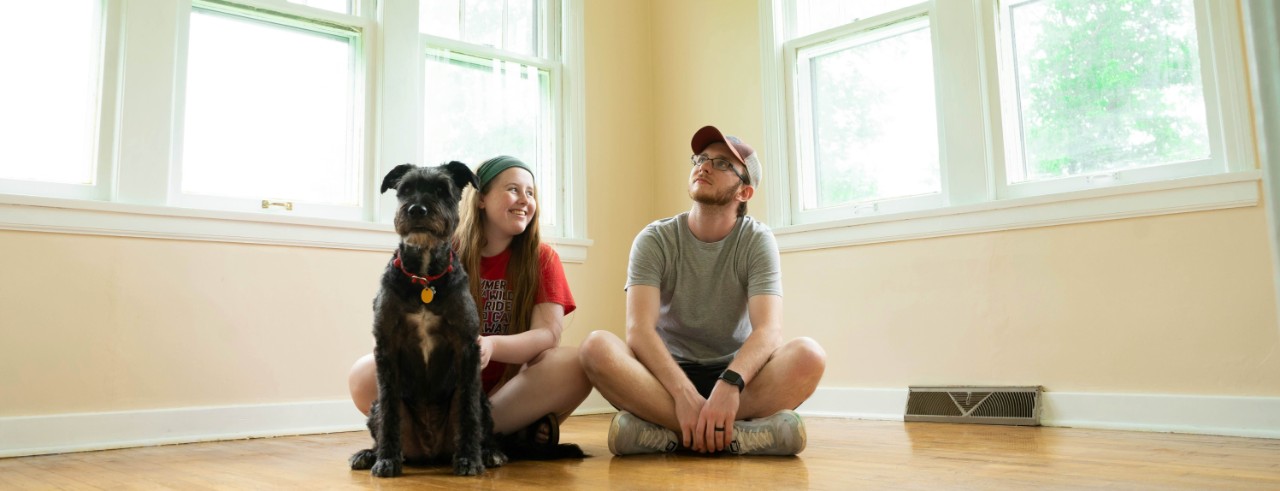 A man, woman and their dog sit inside an empty room of a home they just moved into.